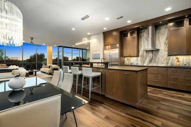 kitchen with dark hardwood / wood-style flooring, stainless steel built in refrigerator, wall chimney range hood, an inviting chandelier, and a center island