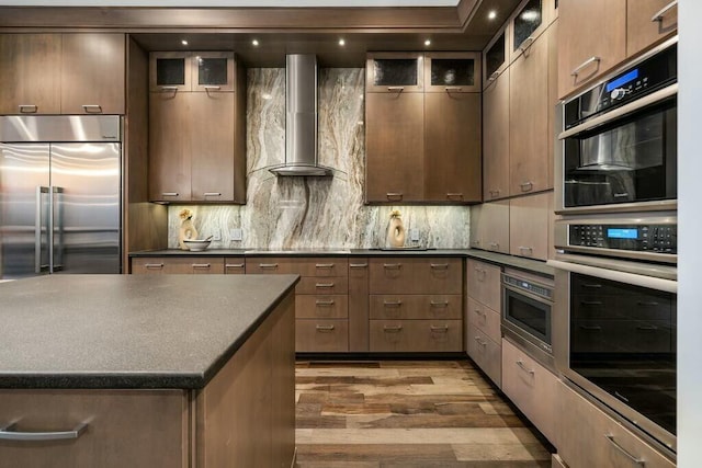 kitchen featuring backsplash, wall chimney exhaust hood, wood-type flooring, and appliances with stainless steel finishes