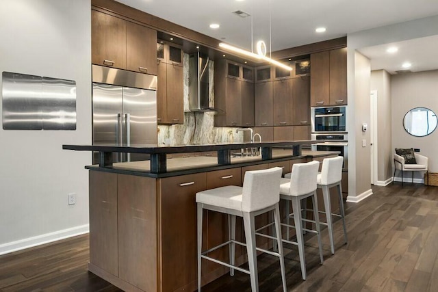 kitchen featuring wall chimney exhaust hood, dark hardwood / wood-style floors, a breakfast bar area, a kitchen island with sink, and appliances with stainless steel finishes