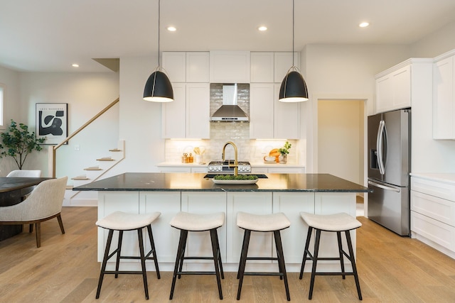 kitchen featuring a center island with sink, wall chimney range hood, decorative light fixtures, white cabinetry, and stainless steel fridge with ice dispenser