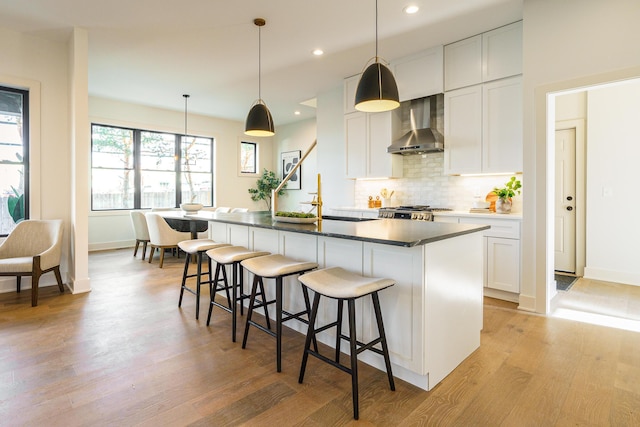 kitchen with wall chimney range hood, hanging light fixtures, white cabinets, and a kitchen island with sink