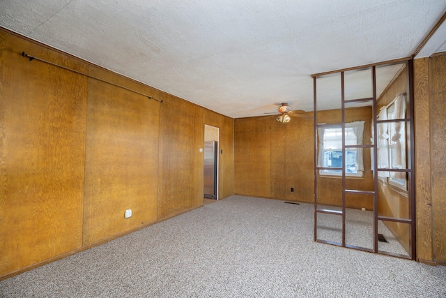 empty room with ceiling fan, a textured ceiling, light carpet, wood walls, and visible vents