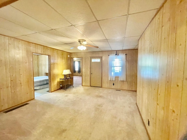 interior space with carpet flooring, a drop ceiling, wooden walls, and ceiling fan