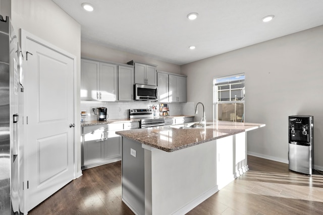 kitchen with light stone countertops, appliances with stainless steel finishes, dark hardwood / wood-style flooring, sink, and a center island with sink