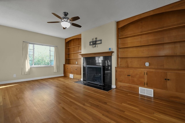 unfurnished living room with ceiling fan, wood-type flooring, built in shelves, and a tiled fireplace