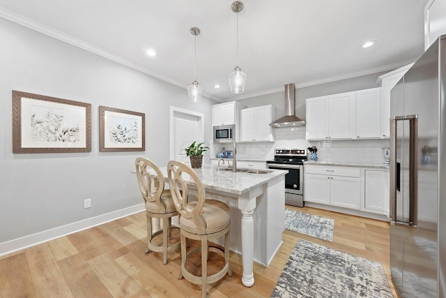 kitchen with white cabinets, wall chimney range hood, light wood-type flooring, an island with sink, and appliances with stainless steel finishes