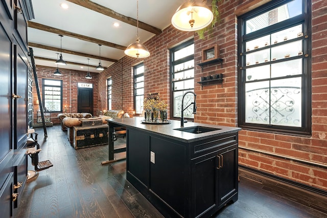 kitchen featuring beam ceiling, sink, hanging light fixtures, brick wall, and an island with sink