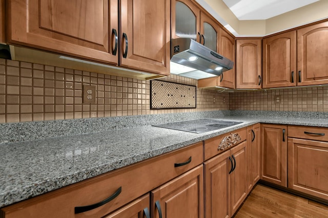 kitchen with wall chimney exhaust hood, light hardwood / wood-style flooring, backsplash, stone countertops, and black electric cooktop