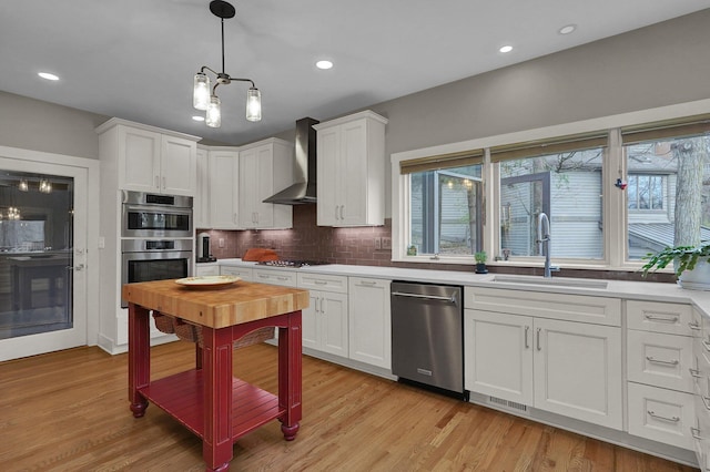 kitchen with wall chimney range hood, sink, stainless steel appliances, white cabinets, and decorative light fixtures