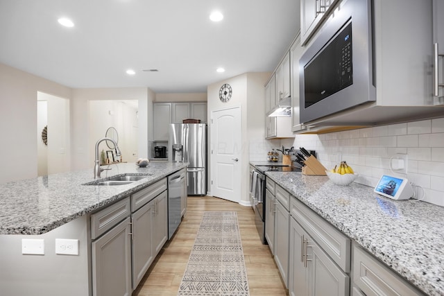 kitchen with sink, light wood-type flooring, an island with sink, light stone counters, and stainless steel appliances