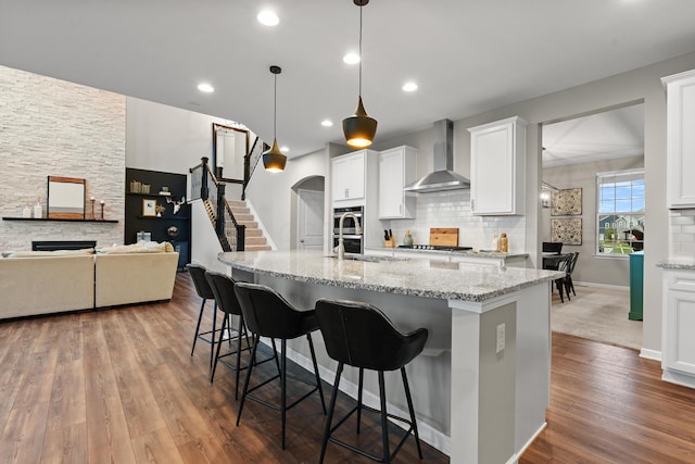 kitchen with a kitchen breakfast bar, wall chimney exhaust hood, pendant lighting, white cabinetry, and an island with sink