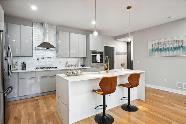 kitchen with wall chimney exhaust hood, hanging light fixtures, an island with sink, appliances with stainless steel finishes, and light wood-type flooring