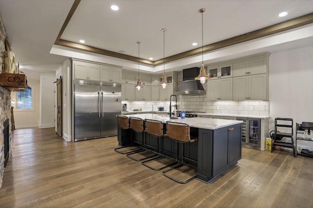 kitchen featuring a kitchen island with sink, dark wood-type flooring, stainless steel built in fridge, hanging light fixtures, and wall chimney exhaust hood