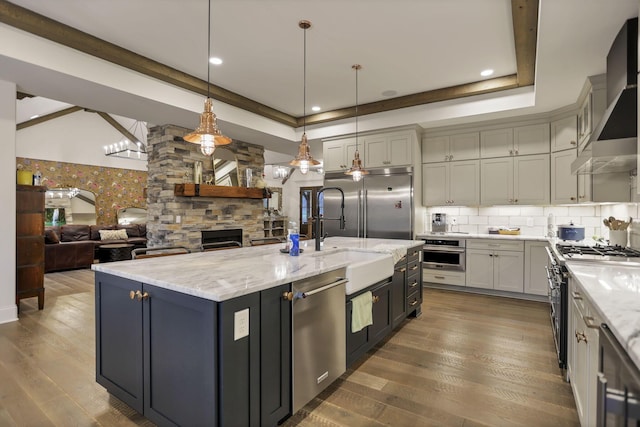 kitchen featuring wall chimney exhaust hood, stainless steel appliances, wood-type flooring, decorative light fixtures, and an island with sink