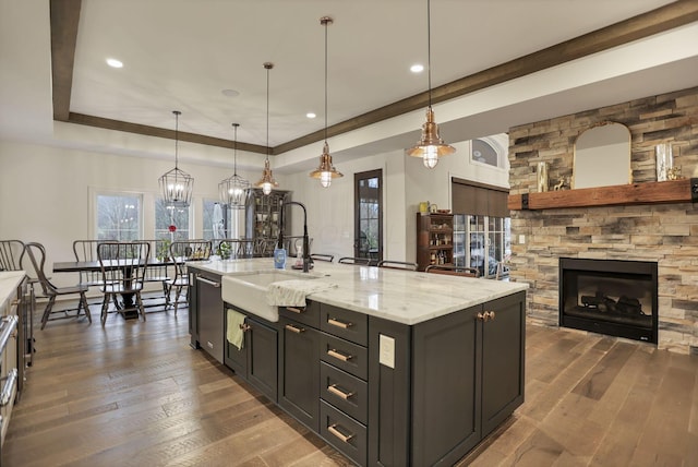 kitchen featuring stainless steel dishwasher, a kitchen island with sink, dark wood-type flooring, a stone fireplace, and hanging light fixtures