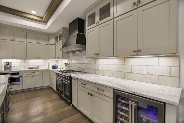 kitchen with dark wood-type flooring, wall chimney range hood, appliances with stainless steel finishes, light stone counters, and beverage cooler