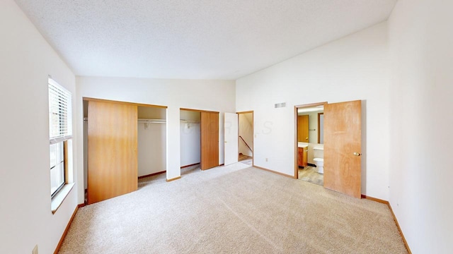 unfurnished bedroom with ensuite bathroom, a textured ceiling, light colored carpet, lofted ceiling, and multiple closets