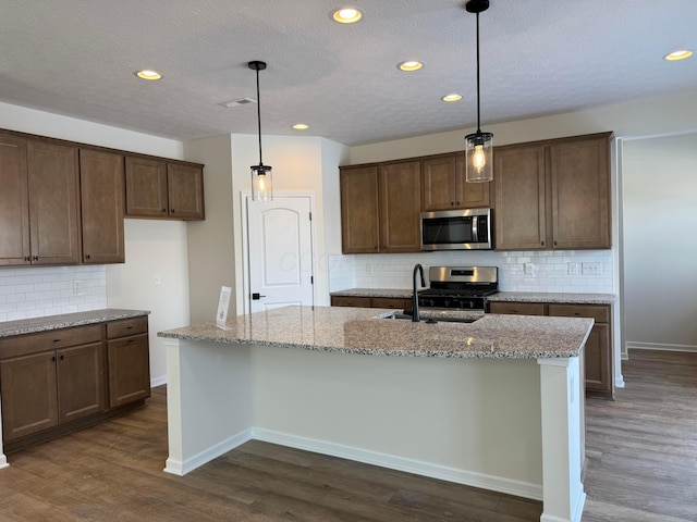 kitchen with a center island with sink, a sink, light stone counters, dark wood finished floors, and appliances with stainless steel finishes