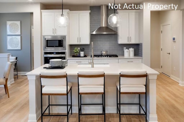 kitchen with wall chimney exhaust hood, stainless steel appliances, an island with sink, and white cabinets