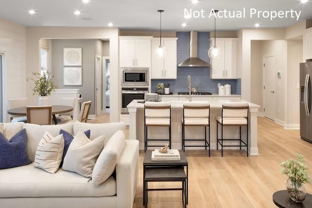 kitchen with wall chimney range hood, decorative light fixtures, appliances with stainless steel finishes, and a center island with sink