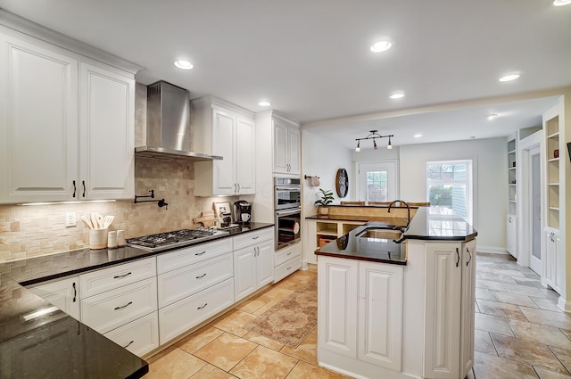 kitchen featuring sink, stainless steel appliances, wall chimney range hood, an island with sink, and white cabinets