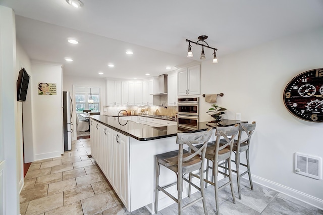 kitchen with wall chimney range hood, kitchen peninsula, tasteful backsplash, a kitchen bar, and white cabinetry