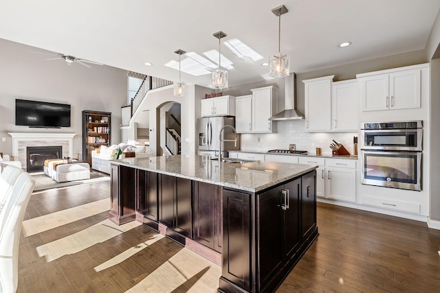 kitchen featuring ceiling fan, a kitchen island with sink, dark wood-type flooring, wall chimney range hood, and decorative light fixtures