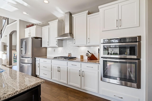 kitchen with appliances with stainless steel finishes, light stone counters, wall chimney range hood, white cabinets, and dark hardwood / wood-style floors