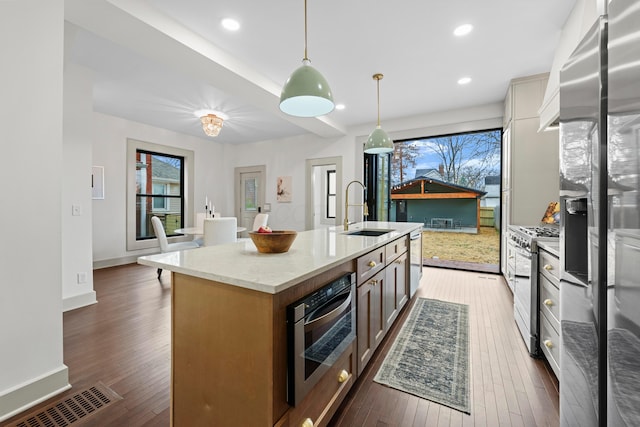 kitchen featuring a kitchen island with sink, dark wood-type flooring, sink, appliances with stainless steel finishes, and decorative light fixtures