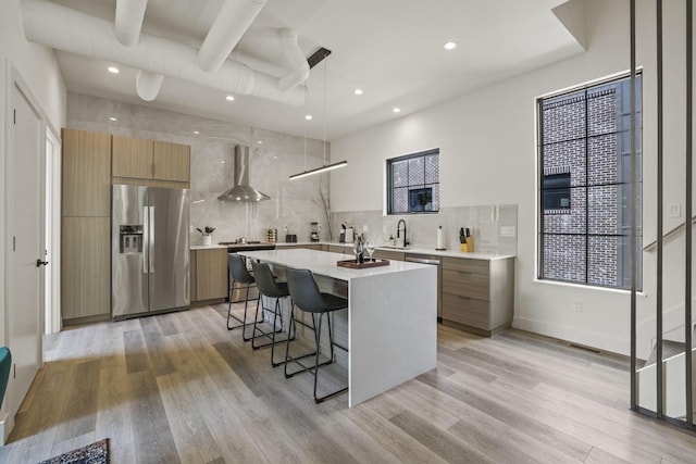 kitchen with stainless steel refrigerator with ice dispenser, light wood-type flooring, hanging light fixtures, and wall chimney exhaust hood