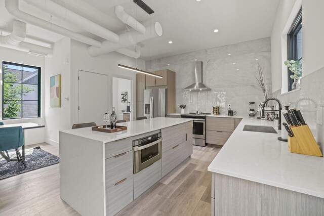 kitchen with stainless steel appliances, sink, wall chimney range hood, light hardwood / wood-style floors, and a kitchen island