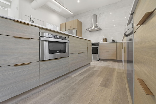 kitchen with appliances with stainless steel finishes, wall chimney exhaust hood, light wood-type flooring, and light brown cabinetry