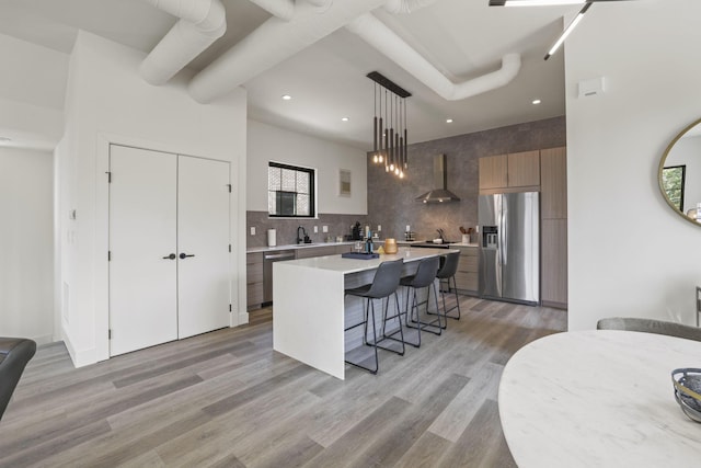 kitchen with wall chimney range hood, hanging light fixtures, light wood-type flooring, a kitchen island, and stainless steel appliances