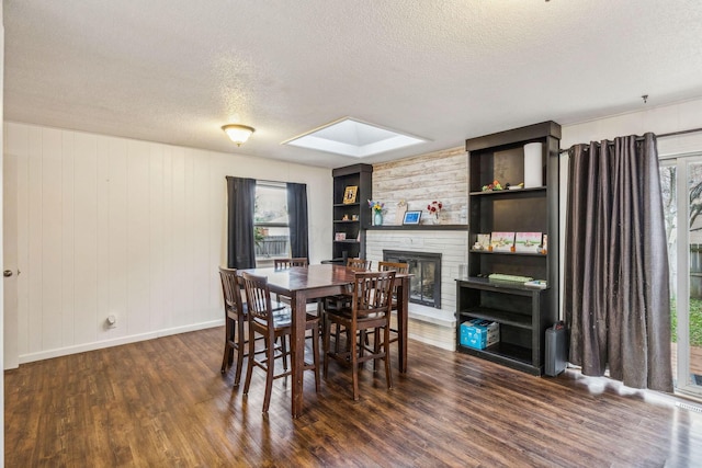 dining room featuring a skylight, dark wood-type flooring, a brick fireplace, wood walls, and a textured ceiling