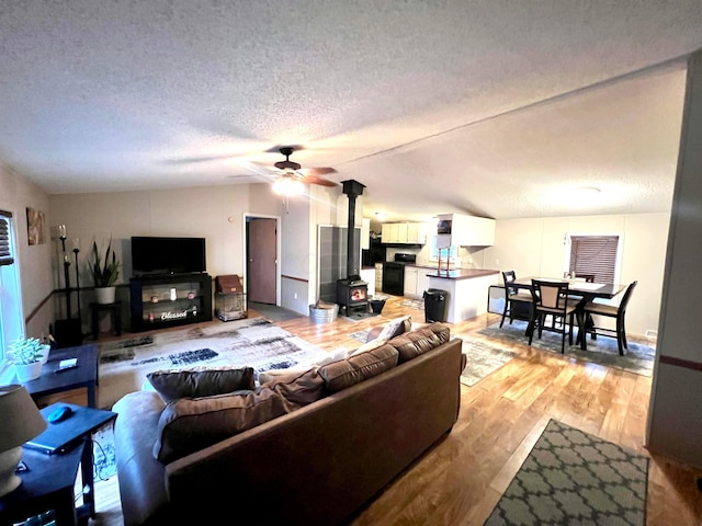 living room with vaulted ceiling, a wood stove, ceiling fan, a textured ceiling, and light hardwood / wood-style flooring