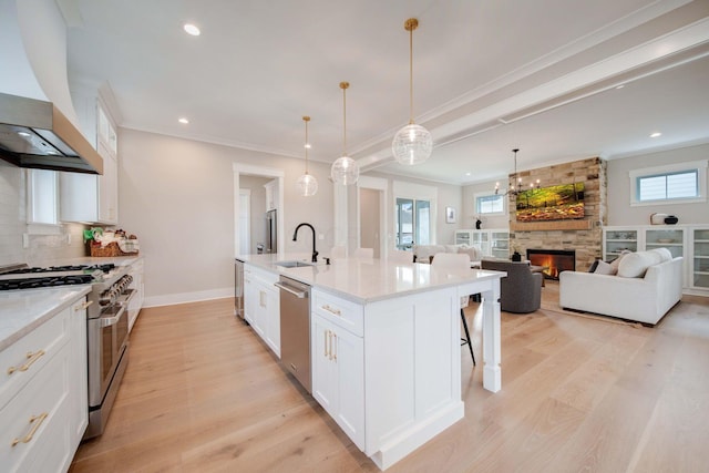 kitchen with custom exhaust hood, a center island with sink, a stone fireplace, white cabinetry, and stainless steel appliances