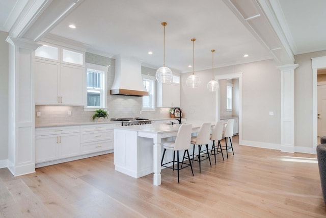 kitchen featuring custom exhaust hood, a kitchen island with sink, crown molding, pendant lighting, and white cabinetry