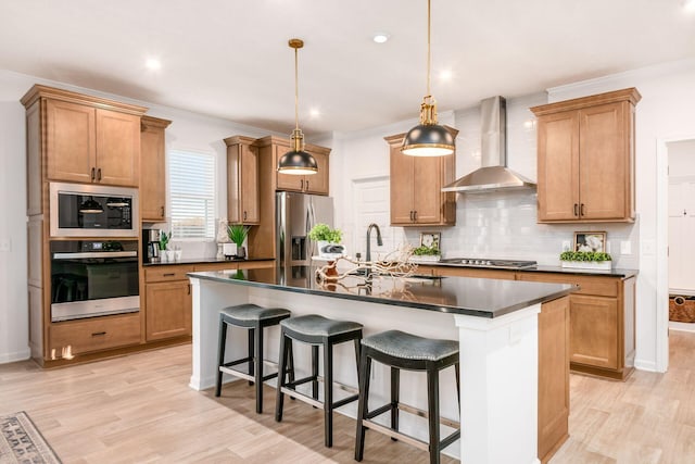 kitchen with wall chimney exhaust hood, hanging light fixtures, stainless steel appliances, a kitchen island with sink, and light wood-type flooring