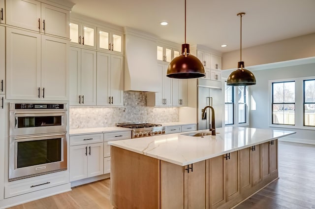 kitchen with a sink, appliances with stainless steel finishes, backsplash, a center island with sink, and custom range hood