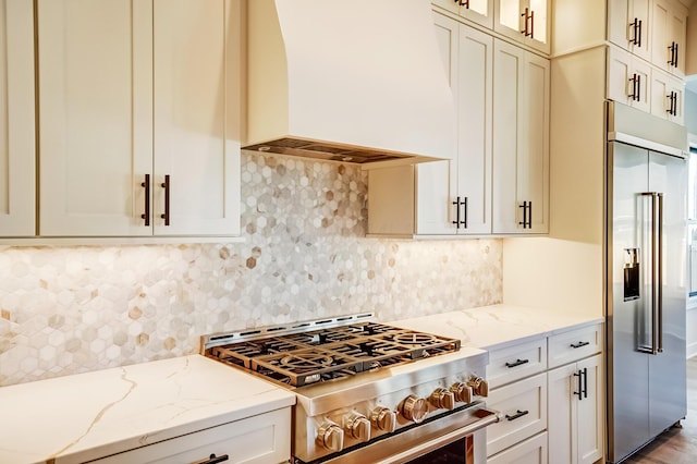 kitchen featuring decorative backsplash, premium appliances, glass insert cabinets, light stone counters, and custom exhaust hood