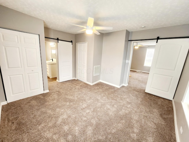 unfurnished bedroom featuring ensuite bath, ceiling fan, a barn door, light colored carpet, and a textured ceiling