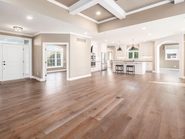 unfurnished living room with sink, ornate columns, light wood-type flooring, ornamental molding, and beam ceiling