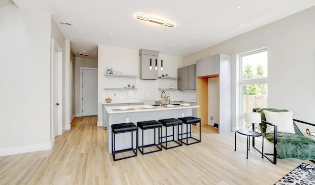 kitchen with gray cabinetry, a kitchen island with sink, tasteful backsplash, a kitchen bar, and light wood-type flooring