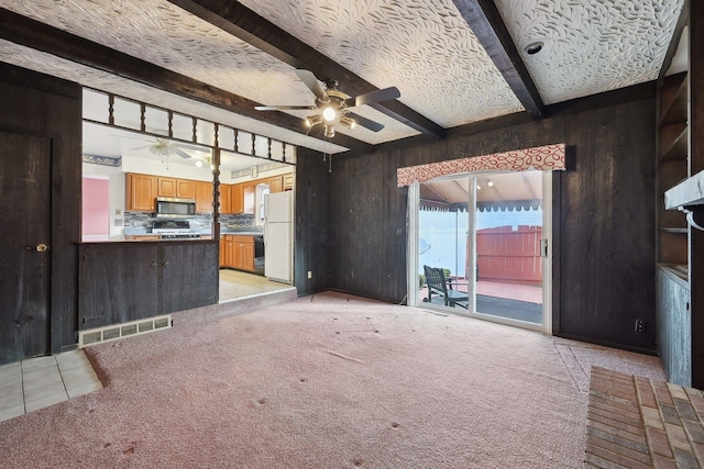 unfurnished living room featuring beamed ceiling, ceiling fan, light colored carpet, and wooden walls