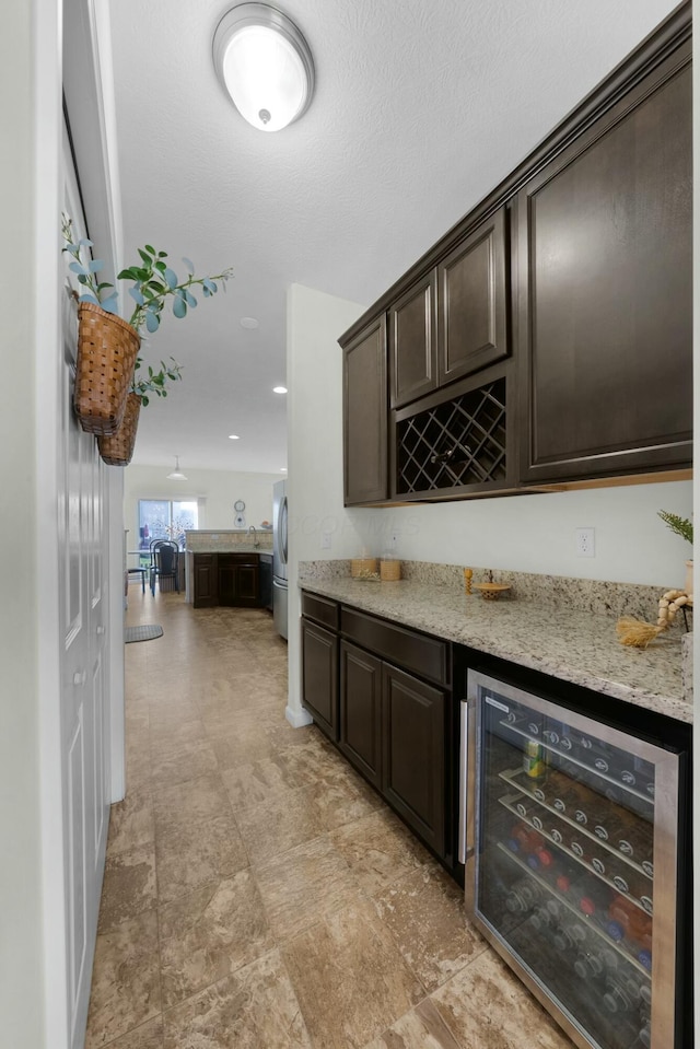 bar featuring stainless steel refrigerator, dark brown cabinetry, light stone countertops, and beverage cooler