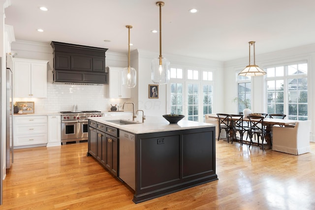 kitchen with sink, a center island with sink, hanging light fixtures, stainless steel appliances, and white cabinets