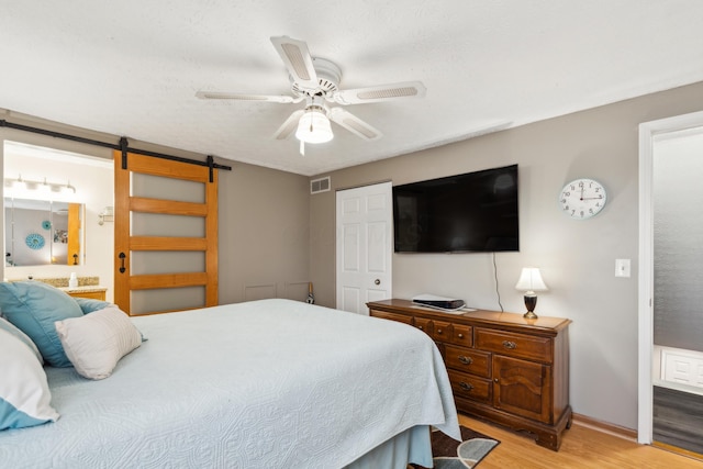 bedroom featuring light wood-type flooring, a textured ceiling, ceiling fan, a barn door, and connected bathroom