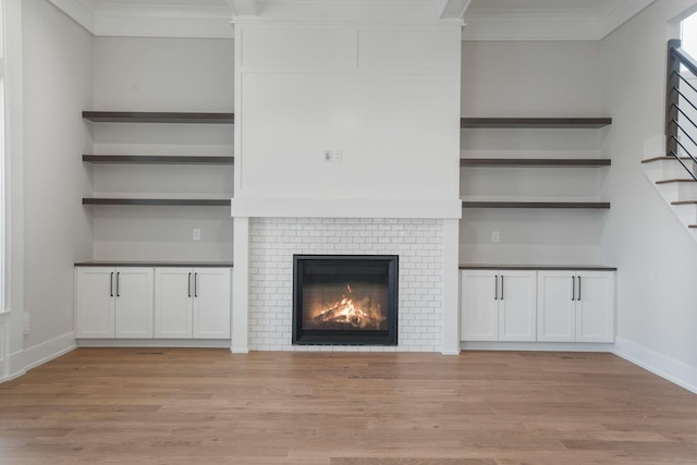 unfurnished living room with a brick fireplace, stairway, light wood-style floors, and crown molding