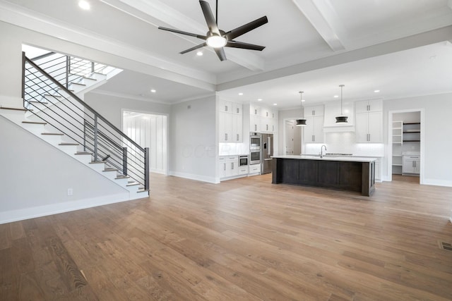 unfurnished living room featuring baseboards, ceiling fan, beamed ceiling, stairs, and light wood-style floors