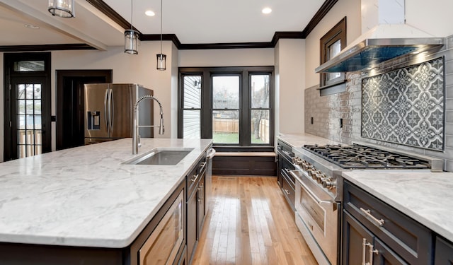 kitchen with a center island with sink, wall chimney range hood, sink, hanging light fixtures, and stainless steel appliances